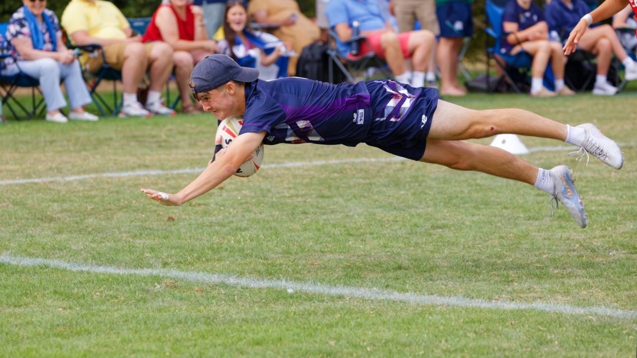 A Scotland player in a cap dives horizontally through the air with the ball, with spectators watching from camping chairs behind