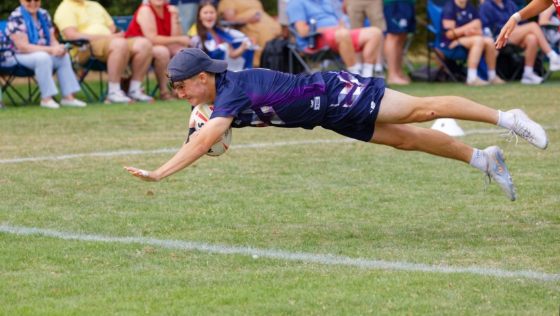 A Scotland player in a cap dives horizontally through the air with the ball, with spectators watching from camping chairs behind