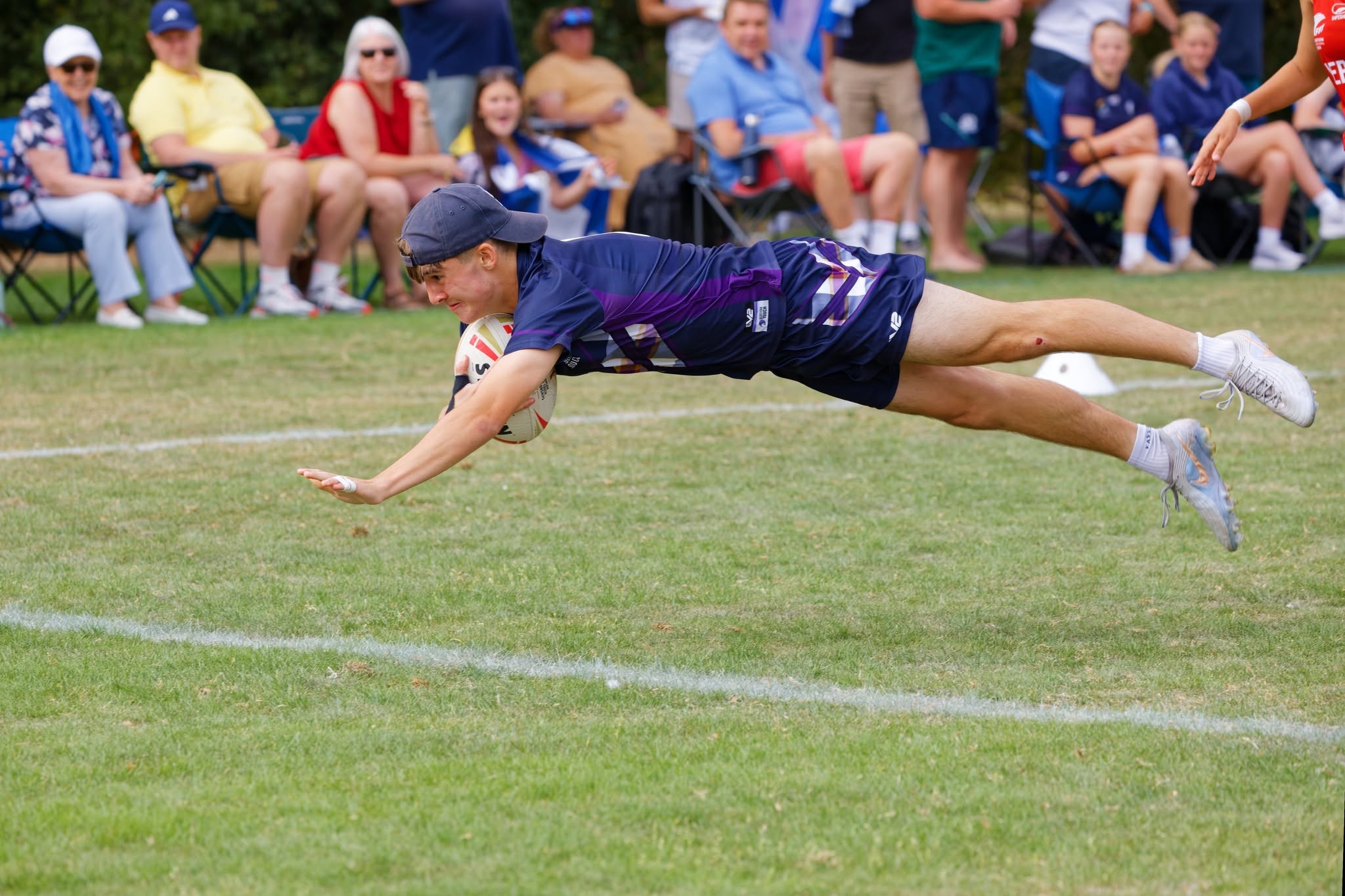 A Scotland player in a cap dives horizontally through the air with the ball, with spectators watching from camping chairs behind