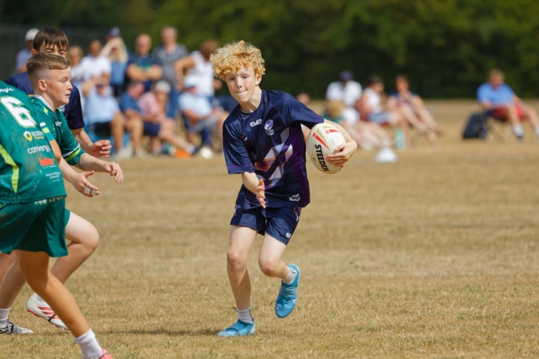 A Scotland player in navy and purple kit carries the ball and prepares to pass as opponents in green close in, with spectators behind