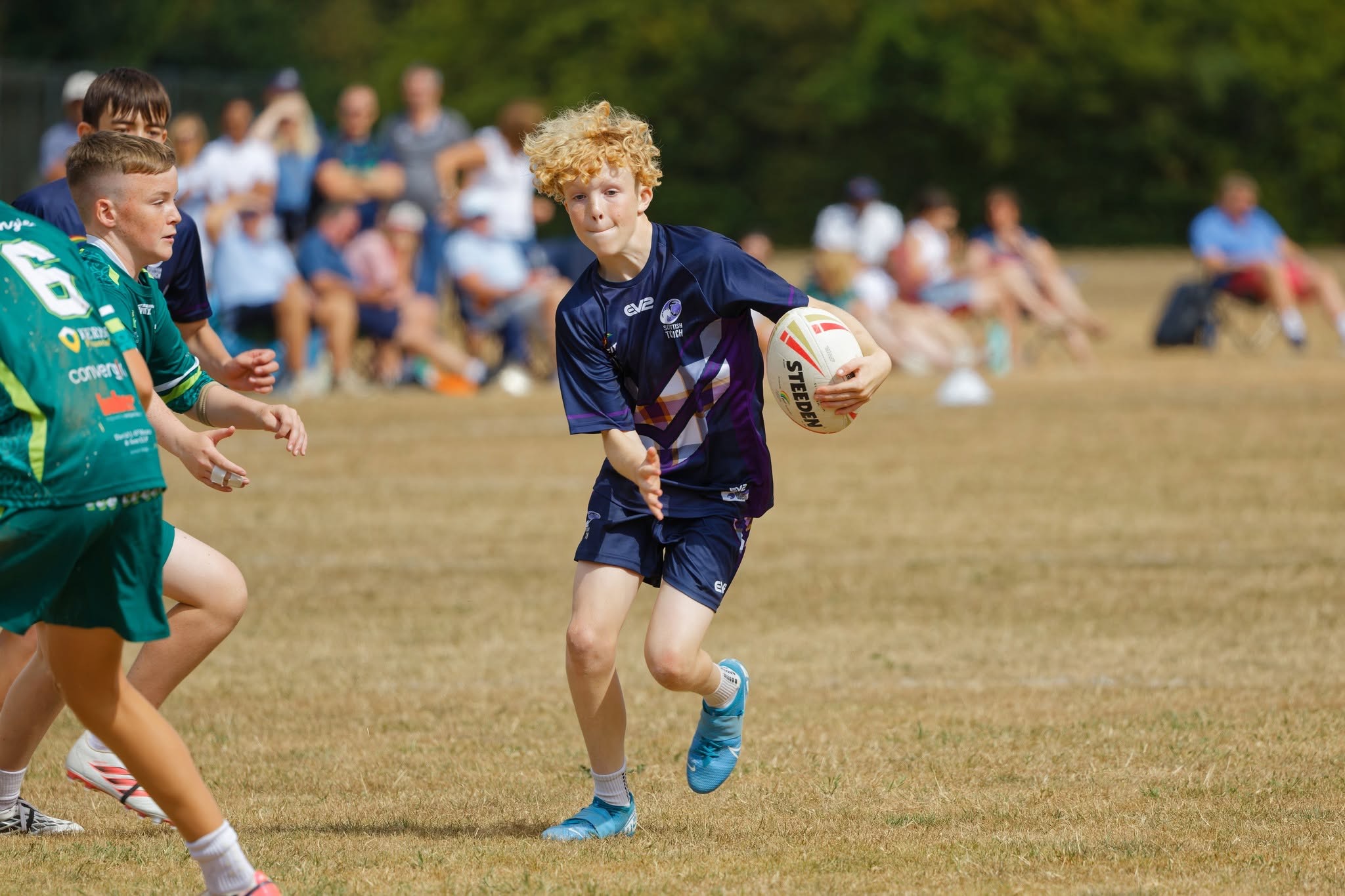 A Scotland player in navy and purple kit carries the ball and prepares to pass as opponents in green close in, with spectators behind
