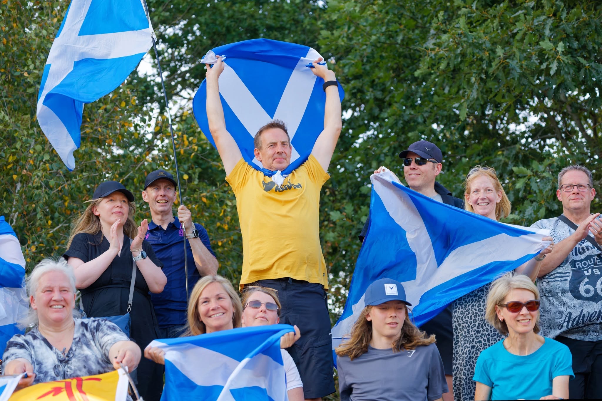 A crowd of Scotland supporters smiling and clapping, with a man in a yellow t-shirt holding a saltire above his head