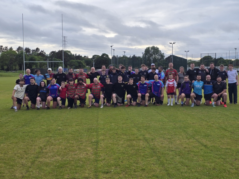 A large group of Dundee Touch players of all ages in mixed kit, some standing and some kneeling, on a grass pitch under a cloudy sky