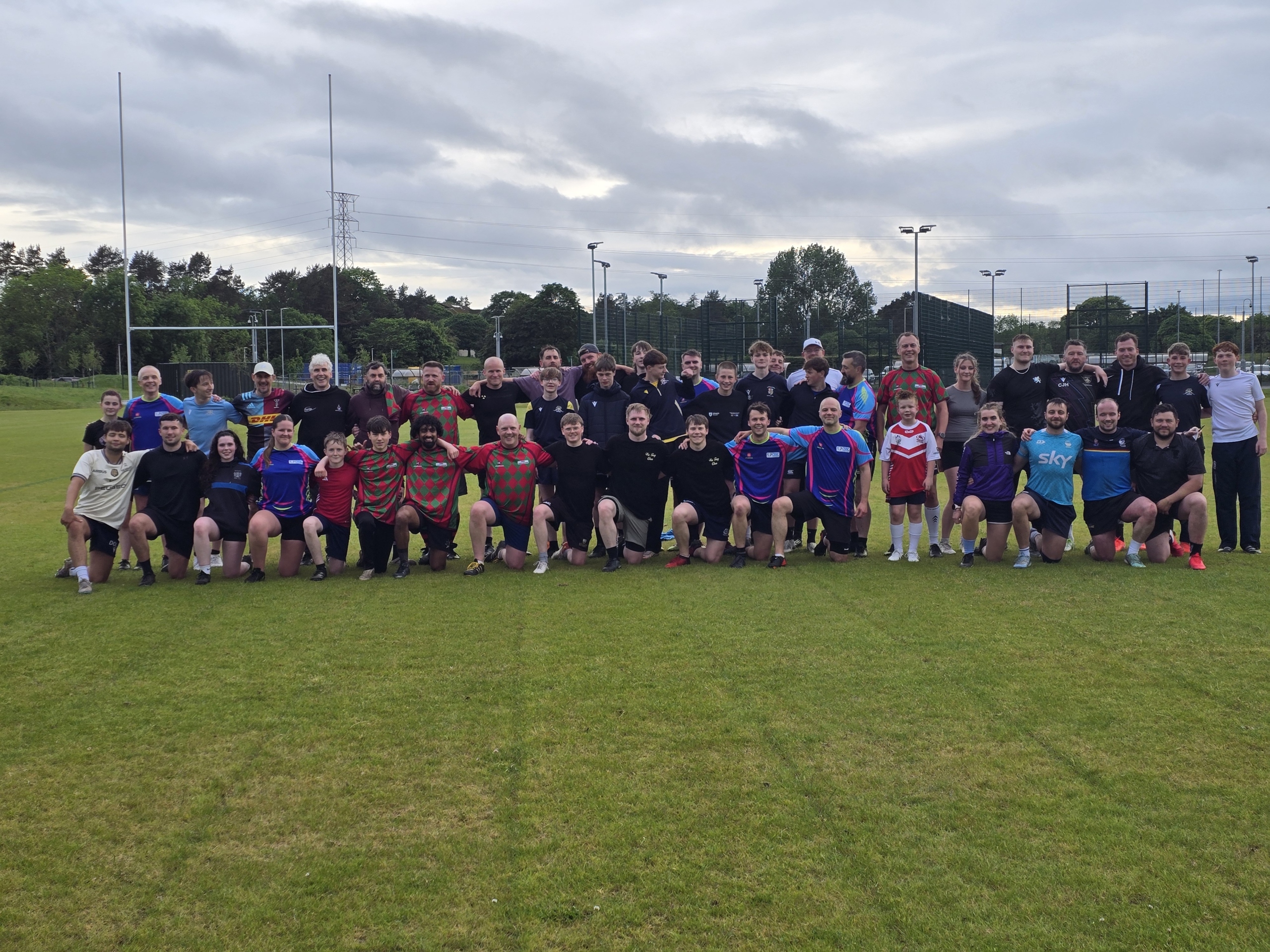 A large group of Dundee Touch players of all ages in mixed kit, some standing and some kneeling, on a grass pitch under a cloudy sky